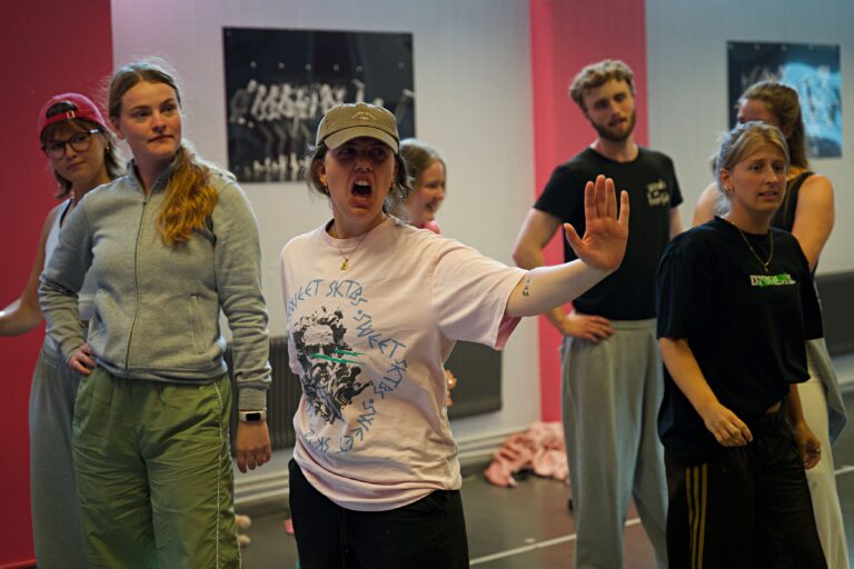 Bild på sju personer som övar pjäsen Legally Blonde. De står alla fyra i träningskläder i en danssal. En av den ser ut att skrika och sätter upp handen som för att säga stopp.