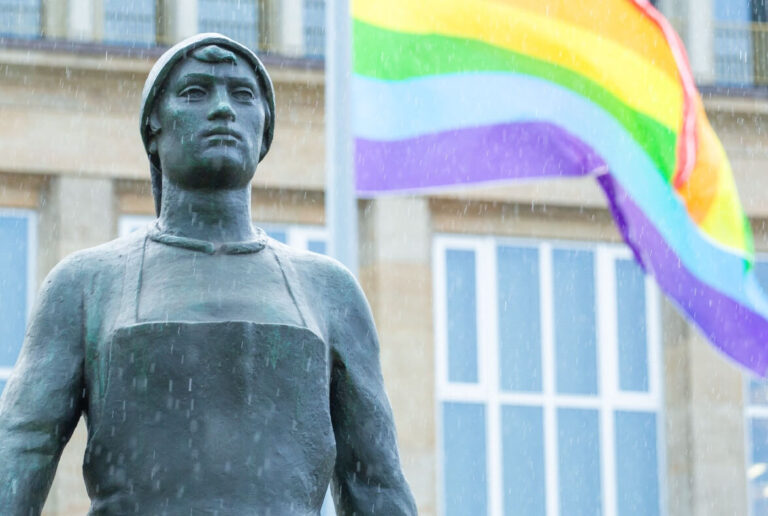 Bild på en staty föreställande en arbetande person och bakom denna ser man regnbågsflaggan. Bilden är tagen i den tyska staden Dresden.