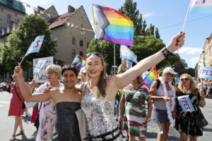 Bild från Stockholms Pride-parad där man ser Rådhuset i bakgrunden, en grupp glada människor med pappersflaggor som det står FPES på.