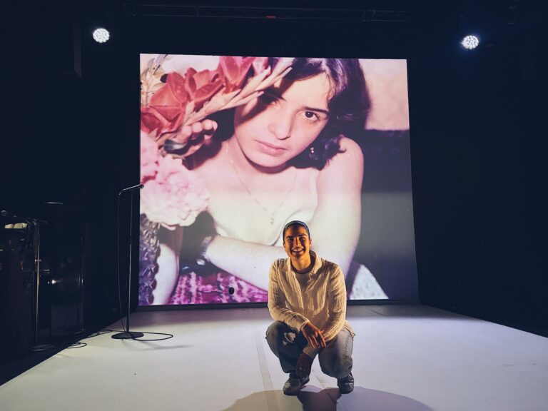 Skådespelaren Beri Gerwise sitter på huk. I bakgrunden finns en bild på hennes mamma som liten projicerad på en skärm. Beri är på scen i en black box-teater.