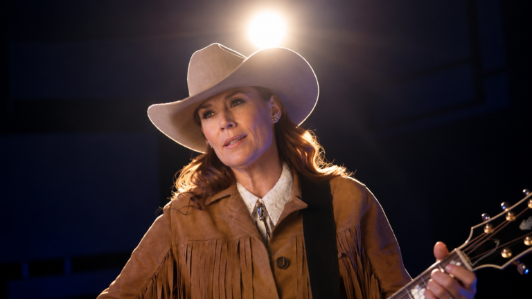 Bild på Jill Johnson med gitarr i hand och cowboyhatt på en scen med strålkastarljus bakom sig.