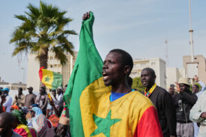 Bild på en man i en demonstration mot homo- bi och transpersoner. Han ropar och håller upp höger arm med knuten näve. Han är draperad i Senegals flagga. Runt honom finns flera männiksor och i bakgrunden ser man ett stort palmträd.