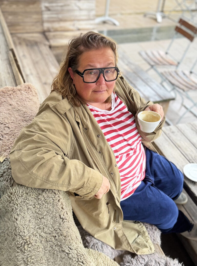 Anna Högkil i glasögon sitter på en bänk på et kafé med en kaffekopp i handen och tittar in i kameran, hon har en uppknäppt jacka och en tröja på sig.