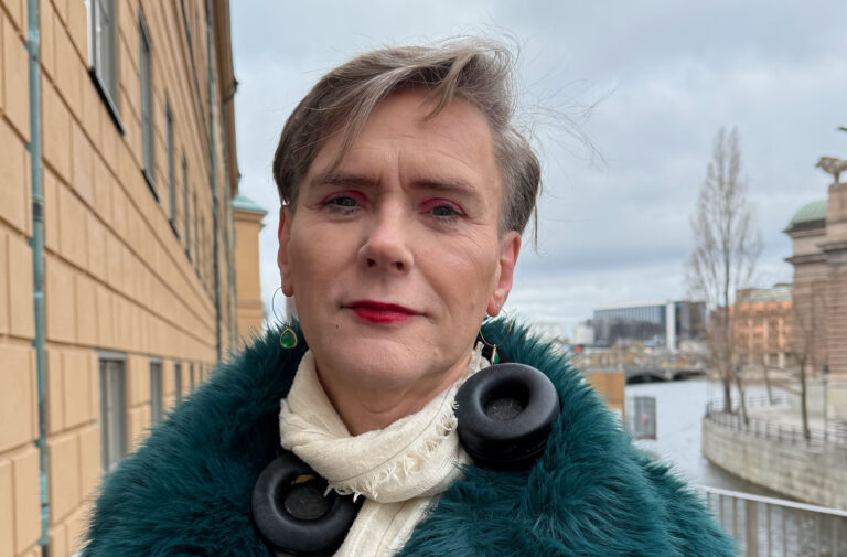 Bild på Helena Berglund stående utanför riksdagshuset i Stockholm i en pälsjacka, halsduk och hörlurar. I bakgrunden syns delar av riksdagshuset och Stockholms Stadshus.