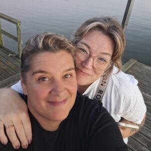 Lenie och Ninna står på en brygga med vatten i bakgrunden. Bilden är en selfie och Ninna har sin högra hand på Lenies högra axel. Lenie tar bilden